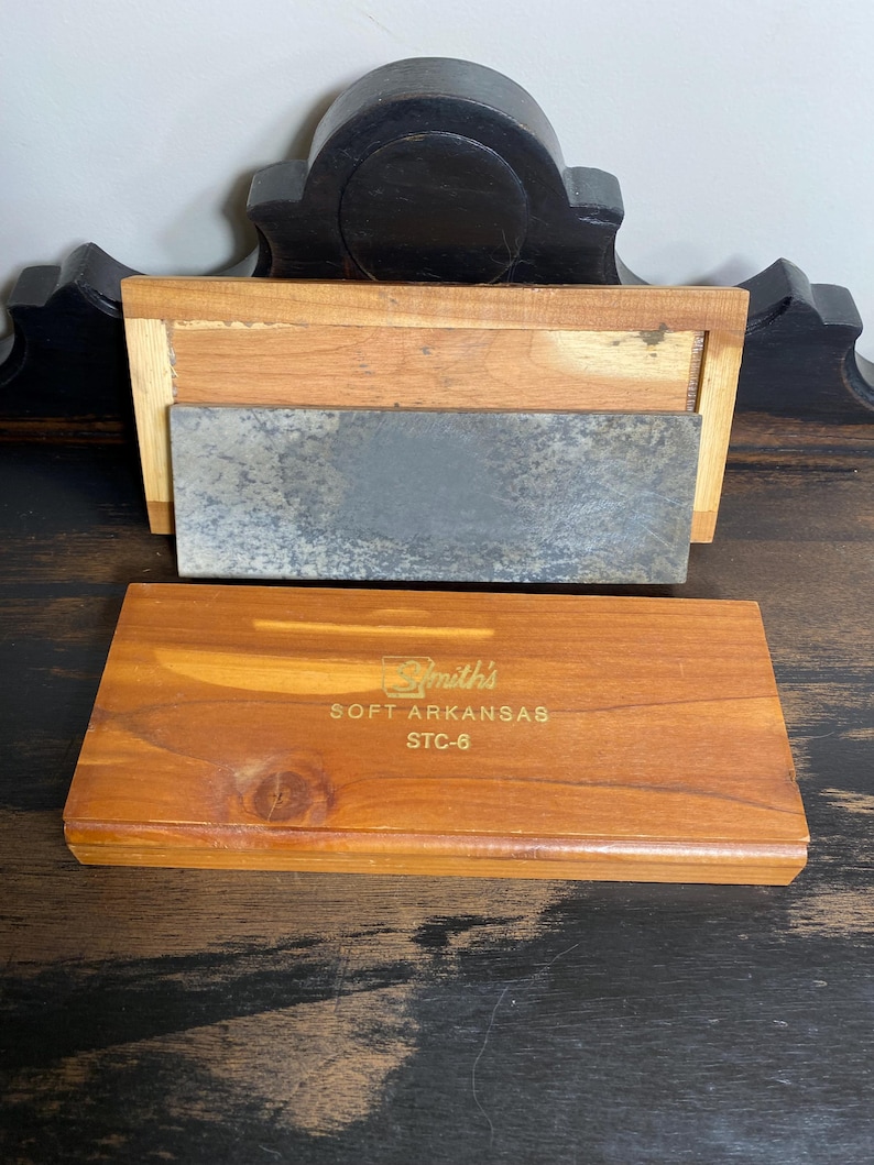 May include: A wooden sharpening stone set with a rectangular, gray stone and a wooden base. The set includes a separate wooden box with the text "Smith's SOFT ARKANSAS STC-6" in gold lettering. The wood has a warm, reddish-brown tone.