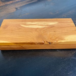 May include: A rectangular wooden box with a natural wood grain finish. The box is a warm, light brown color and has a slightly rough edge on one side. The lid appears to be hinged, suggesting it opens to reveal storage space.