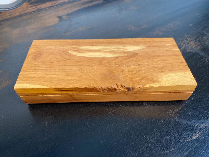 May include: A rectangular wooden box with a natural wood grain finish. The box is a warm, light brown color and has a slightly rough edge on one side. The lid appears to be hinged, suggesting it opens to reveal storage space.