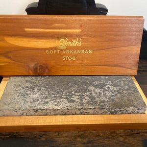 May include: A wooden box with a sliding lid, containing a rectangular sharpening stone. The lid is engraved with "Smith's SOFT ARKANSAS STC-6". The box and stone are set on a dark wooden surface.
