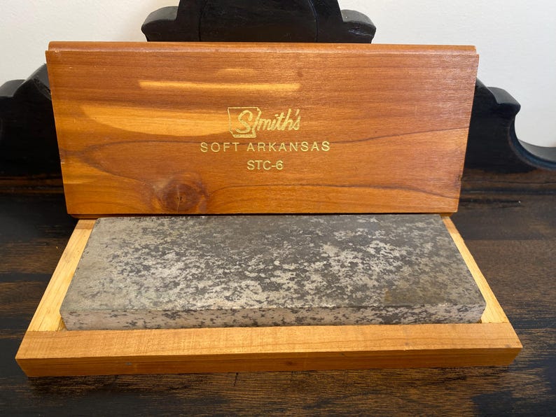 May include: A wooden box with a sliding lid, containing a rectangular sharpening stone. The lid is engraved with "Smith's SOFT ARKANSAS STC-6". The box and stone are set on a dark wooden surface.