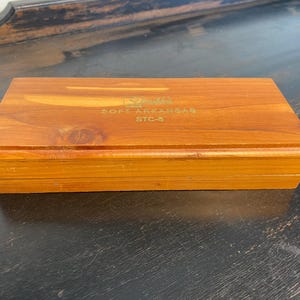 May include: A rectangular wooden box with a warm brown finish. The box has a hinged lid and is labeled with gold text that reads "SOFT ARKANSAS STC-6". The box is sitting on a dark surface.