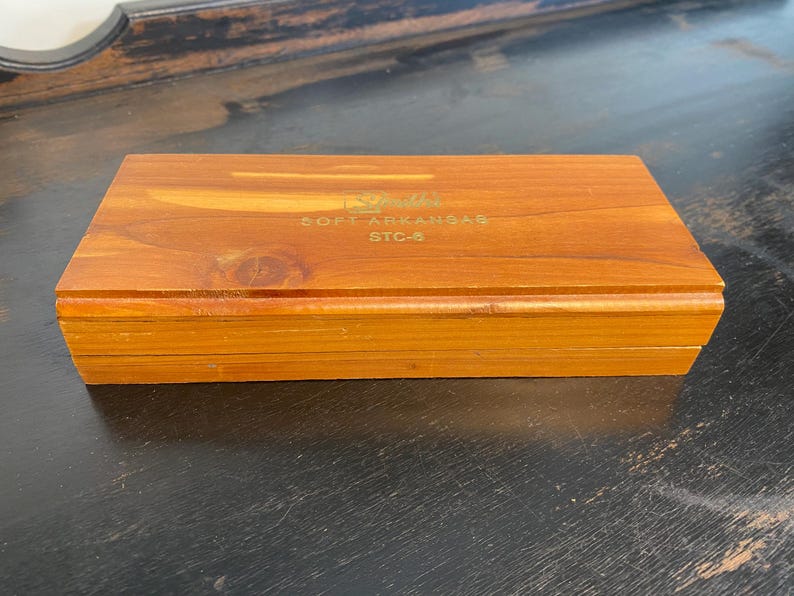 May include: A rectangular wooden box with a warm brown finish. The box has a hinged lid and is labeled with gold text that reads "SOFT ARKANSAS STC-6". The box is sitting on a dark surface.