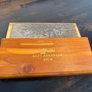 May include: A wooden box with a sliding lid, containing a rectangular sharpening stone. The box is made of light brown wood and has the text "Smith's SOFT ARKANSAS STC-6" printed on the front. The stone is a gray color.