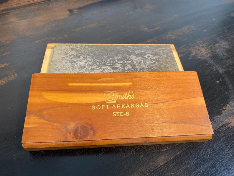 May include: A wooden box with a sliding lid, containing a rectangular sharpening stone. The box is made of light brown wood and has the text "Smith's SOFT ARKANSAS STC-6" printed on the front. The stone is a gray color.