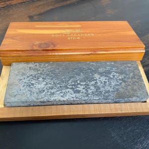 May include: A vintage sharpening stone set. The set includes a rectangular gray stone in a wooden tray, with a wooden lid. The lid is engraved with the text "Smith's Soft Arkansas STC-6". The wood has a warm, reddish-brown tone.