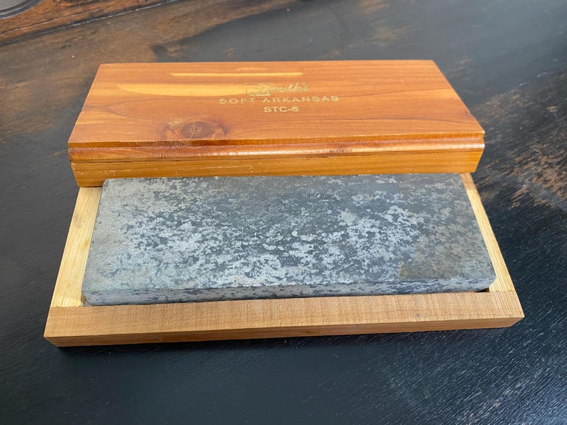 May include: A vintage sharpening stone set. The set includes a rectangular gray stone in a wooden tray, with a wooden lid. The lid is engraved with the text "Smith's Soft Arkansas STC-6". The wood has a warm, reddish-brown tone.