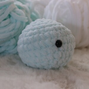 May include: A light blue crocheted stuffed animal with a black button eye. The animal is sitting on a white fluffy surface.