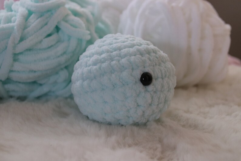 May include: A light blue crocheted stuffed animal with a black button eye. The animal is sitting on a white fluffy surface.