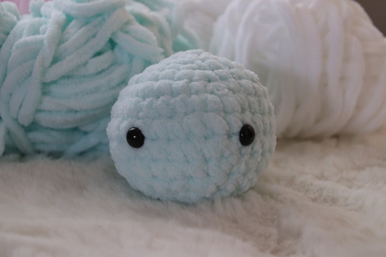 May include: A light blue crocheted stuffed animal with black button eyes. The animal is sitting on a white fluffy surface.