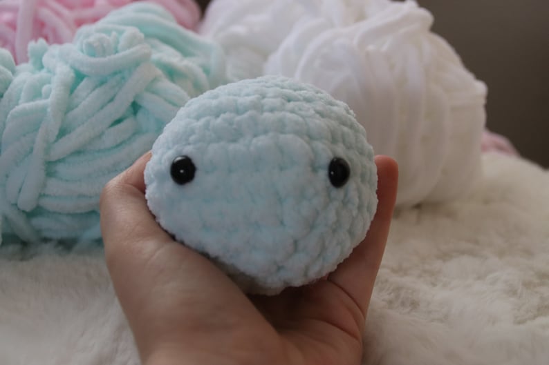 May include: A light blue crocheted stuffed animal with black eyes. The animal is being held in a hand.