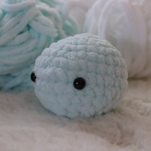 May include: A light blue crocheted stuffed animal with black eyes. The animal is sitting on a white fluffy surface.