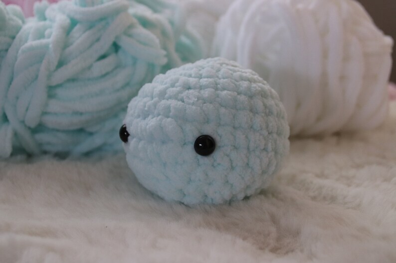 May include: A light blue crocheted stuffed animal with black eyes. The animal is sitting on a white fluffy surface.
