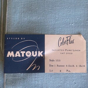 May include: A light blue linen fabric set with a rectangular product tag. The tag reads: STYLED BY MATOUK, ColorFlax, IMPORTED PURE LINEN, VAT DYED, Style 1015, Size 1 Runner 4-12x18, 4 16x16, Set 9 Pcs. The fabric has a visible woven texture.