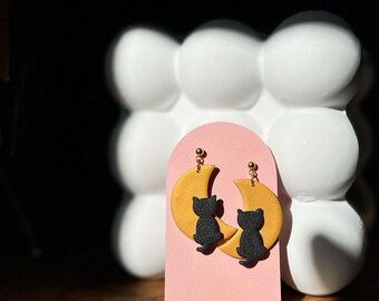 May include: A pair of gold crescent moon earrings with black cat silhouettes. The earrings are on a pink card with the text "GRACE & LILIKO'I".