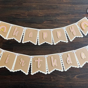 May include: A brown burlap banner with white scalloped edges and pink and gold letters spelling "Happy Birthday". The banner is strung with white ribbon and features a small rainbow with a yellow sun on each end.