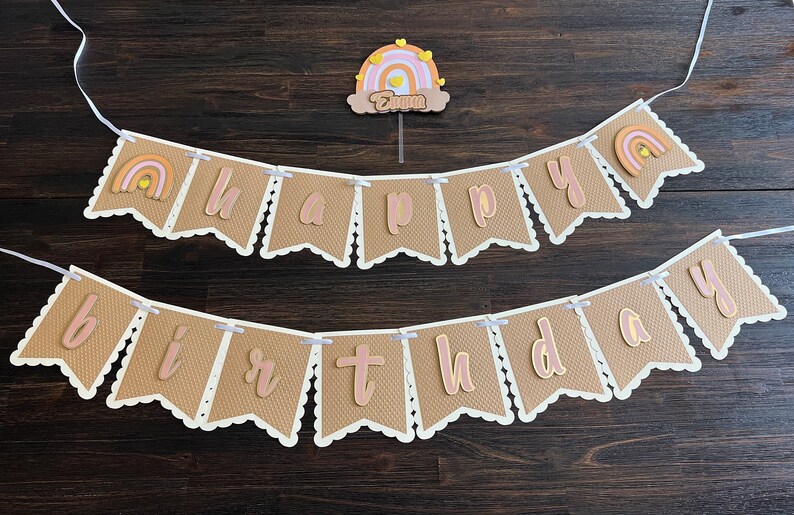 May include: A brown burlap banner with white scalloped edges and pink letters spelling "happy birthday". The banner is strung with white ribbon and features a small rainbow with yellow stars on each flag.