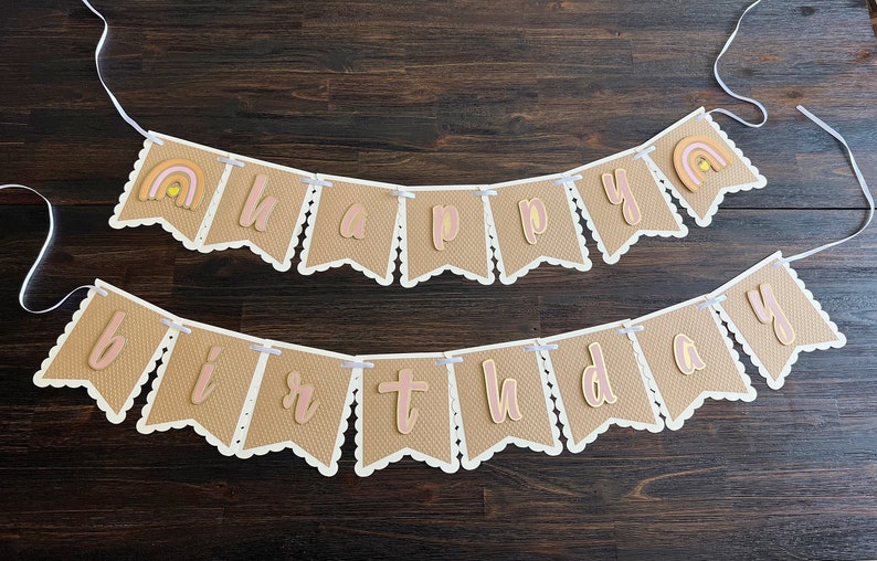 May include: A brown burlap banner with white scalloped edges and pink and gold letters spelling "Happy Birthday". The banner is strung with white ribbon and features a small rainbow with a yellow sun on each end.