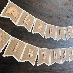 May include: A brown and white banner with the words "happy birthday" in pink and gold letters. The banner has a scalloped edge and is decorated with small rainbows.