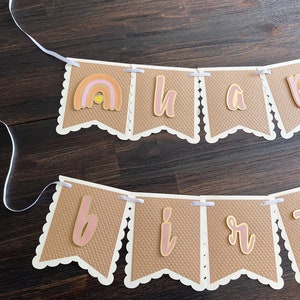 May include: A brown and white banner with the words "Happy Birthday" in pink and gold letters. The banner has a rainbow with a yellow sun in the centre.