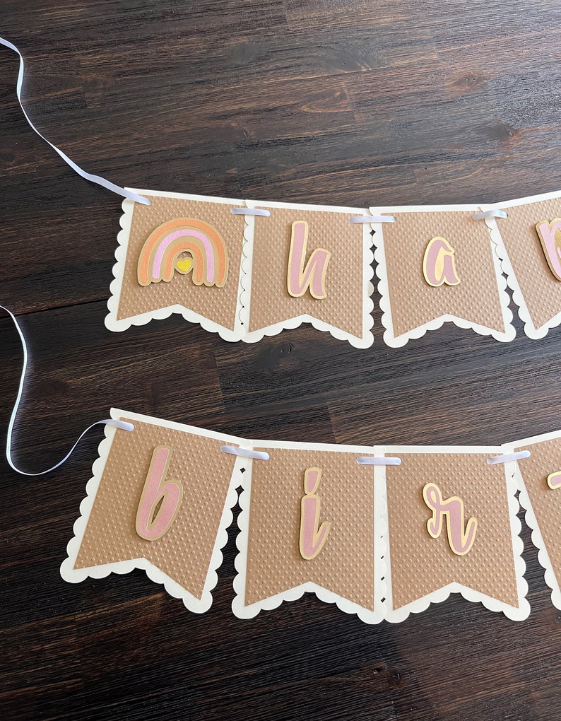 May include: A brown and white banner with the words "Happy Birthday" in pink and gold letters. The banner has a rainbow with a yellow sun in the centre.