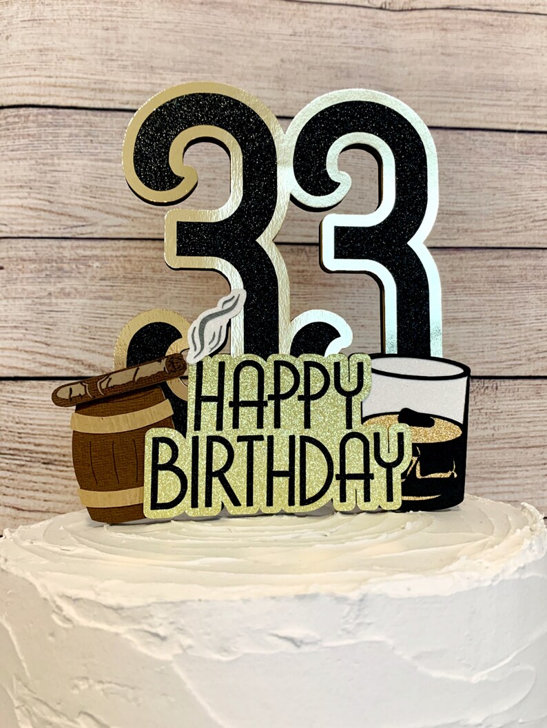 May include: Black and gold glitter cake topper with the number 33, a barrel with a cigar, a glass of whiskey, and the words "Happy Birthday".