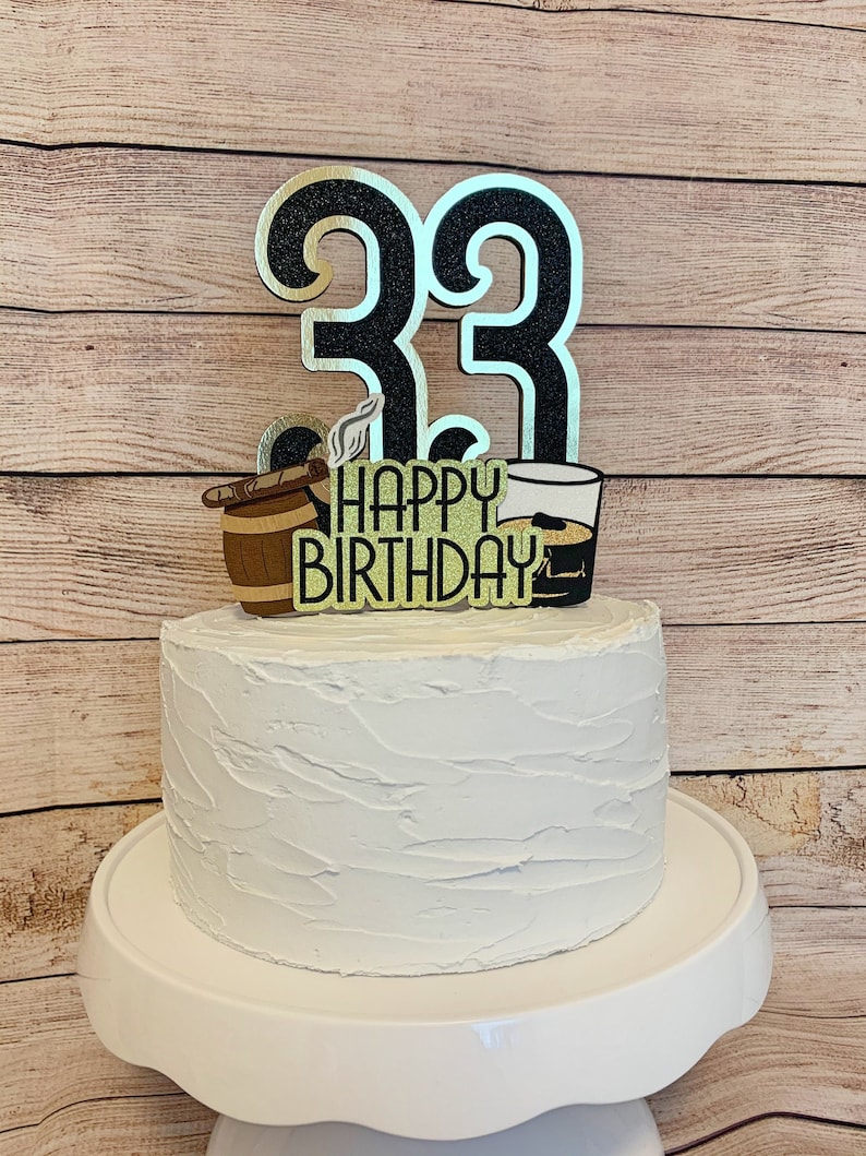 May include: A white cake with black and gold glitter number 33 topper. The topper also includes a cigar, barrel, and a glass of whiskey. The cake is on a white cake stand. The text on the topper reads "Happy Birthday".