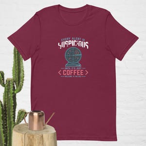May include: A maroon short-sleeve t-shirt featuring a graphic of a radar screen with a coffee cup icon. The text reads: EVERY ALERT IS SUSPICIOUS UNTIL IT'S JUST COFFEE WELCOME TO THE SOC. The design uses light blue, white, and pink colors.