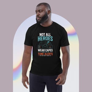 May include: A black short-sleeved t-shirt featuring a graphic print with the text "NOT ALL HEROES WEAR CAPES SOME OF US TUNE ALERTS SO YOU DON'T HAVE TO" in white, light blue, and orange, along with an illustration of a person wearing a headset.