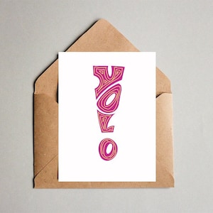 Könnte beinhalten: Eine weiße Karte mit dem Wort "YOLO" in Pink und Gold, in einem braunen Umschlag. Die Buchstaben sind mit einem wellenförmigen Muster stilisiert. Der Umschlag besteht aus braunem Papier und ist teilweise geöffnet, wodurch die Karte sichtbar wird.