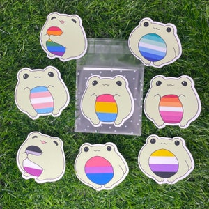 May include: Set of 7 frog stickers featuring different pride flags. Each frog is holding a circle with a different pride flag design. The flags include the transgender, lesbian, bisexual, pansexual, genderqueer, asexual, and non-binary flags.