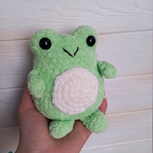 Op de afbeelding: Een groene gehaakte knuffel van een kikker met een witte buik en zwarte ogen.