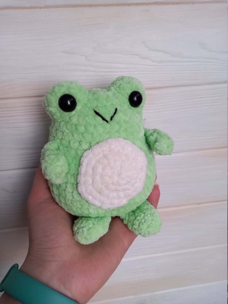 Op de afbeelding: Een groene gehaakte knuffel van een kikker met een witte buik en zwarte ogen.