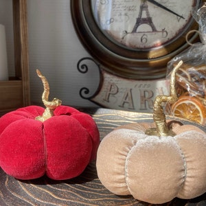 May include: Two decorative velvet pumpkins, one red and one beige, with gold stems. The pumpkins are sitting on a wooden surface.