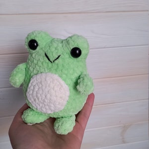 Op de afbeelding: Een groene gehaakte knuffel van een kikker met witte accenten en zwarte ogen. De kikker zit in een hand.