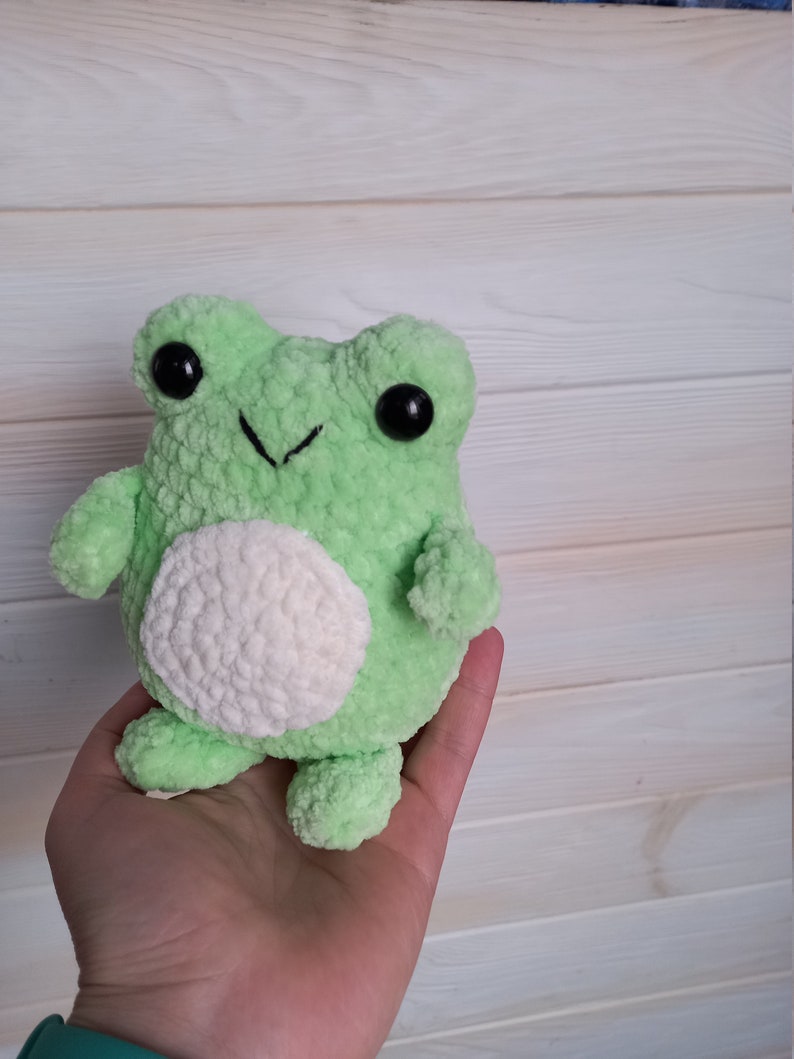 Op de afbeelding: Een groene gehaakte knuffel van een kikker met witte accenten en zwarte ogen. De kikker zit in een hand.