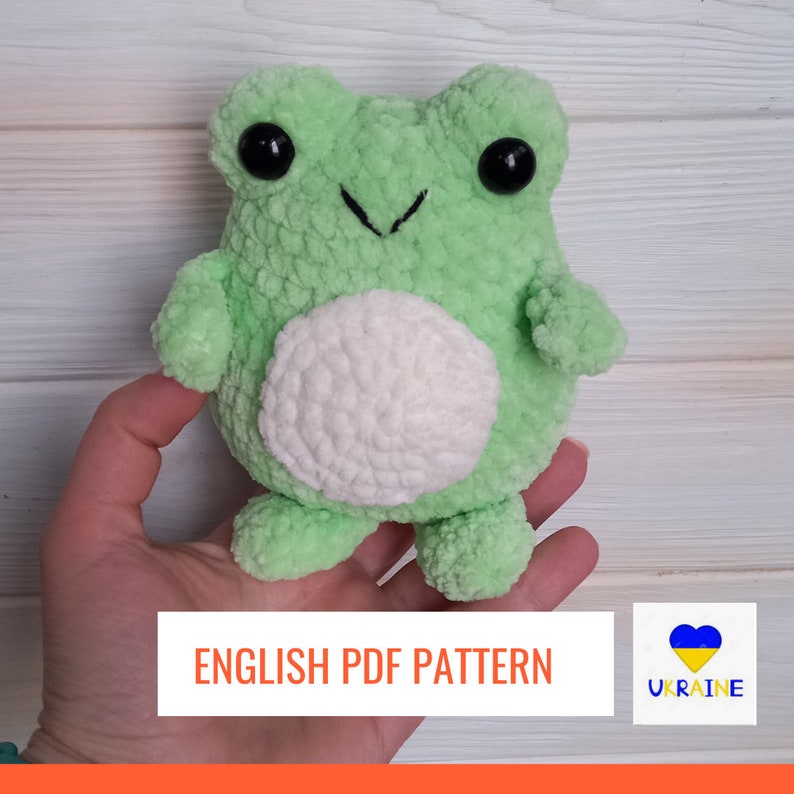 Op de afbeelding: Een gehaakt groen knuffelbeestje in de vorm van een kikker met een witte buik. Het speelgoed wordt in een hand gehouden. De tekst "ENGLISH PDF PATTERN" staat onderaan de afbeelding. Een blauw en geel hart met het woord "UKRAINE" staat rechtsonder.