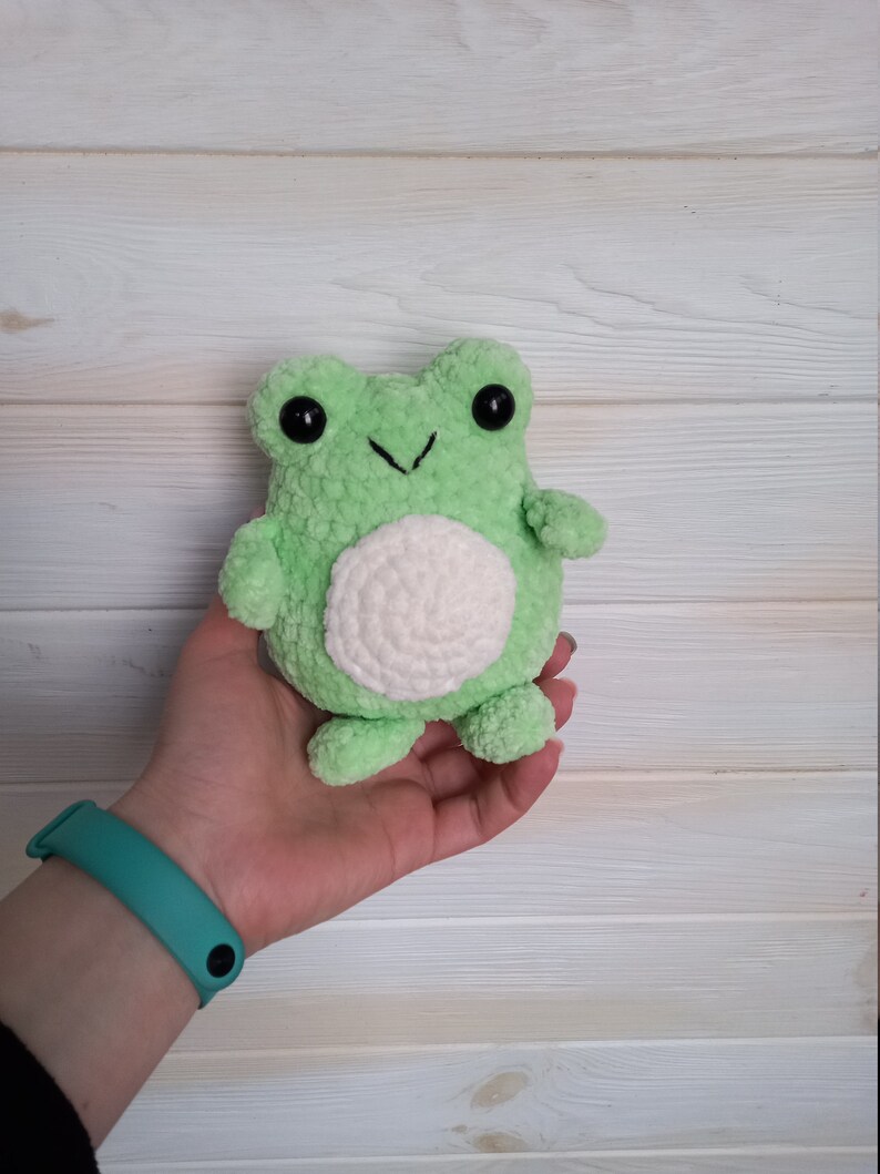 Op de afbeelding: Een groene gehaakte knuffel van een kikker met witte accenten en zwarte ogen. De kikker wordt in een hand vastgehouden.