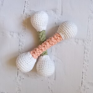 May include: Two crocheted white dumbbells with pink and green accents. The dumbbells are crossed over each other.