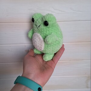 Op de afbeelding: Groene gehaakte kikker knuffel met een witte buik en zwarte ogen. De kikker wordt vastgehouden in een hand met een turquoise polsbandje.