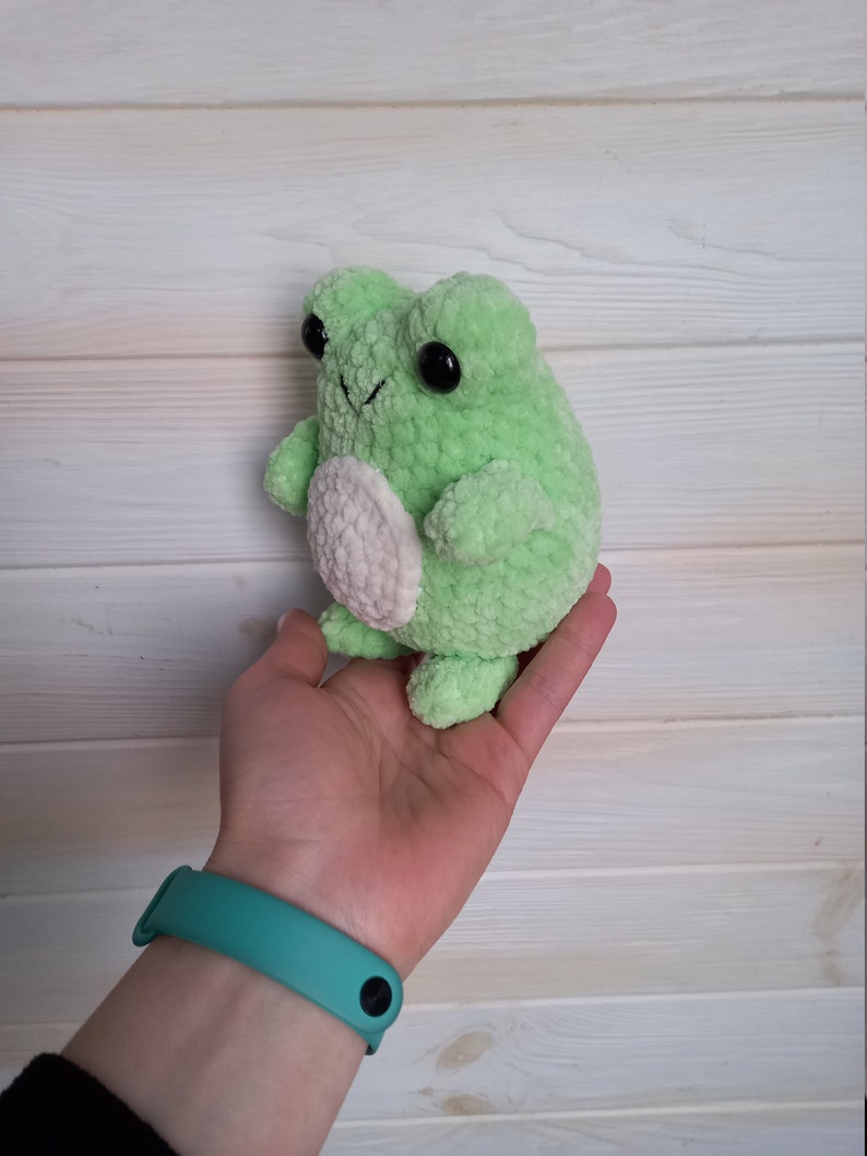 Op de afbeelding: Groene gehaakte kikker knuffel met een witte buik en zwarte ogen. De kikker wordt vastgehouden in een hand met een turquoise polsbandje.