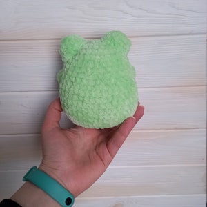 Op de afbeelding: Een klein, groen gehaakt knuffeldier in de vorm van een kikker. De kikker wordt in de hand van een persoon gehouden.