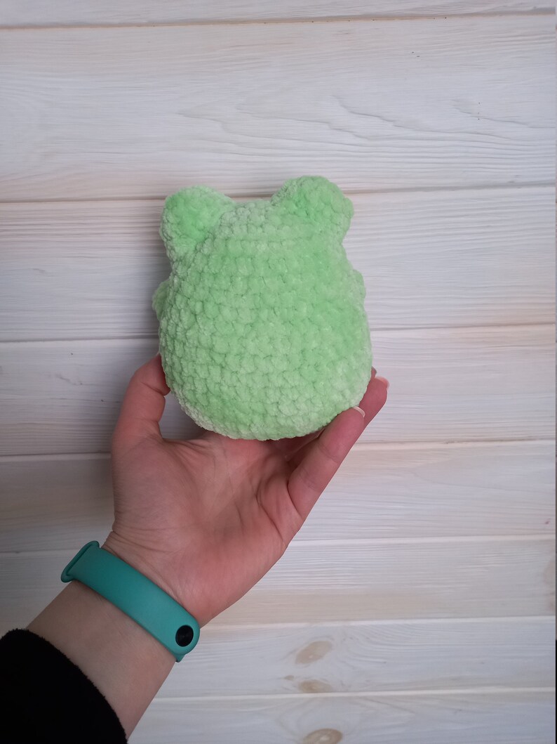 Op de afbeelding: Een klein, groen gehaakt knuffeldier in de vorm van een kikker. De kikker wordt in de hand van een persoon gehouden.
