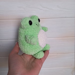 Op de afbeelding: Een groene gehaakte knuffel van een kikker met witte accenten. De kikker heeft zwarte ogen en een kleine, witte buik.