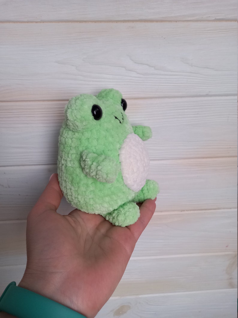Op de afbeelding: Een groene gehaakte knuffel van een kikker met witte accenten. De kikker heeft zwarte ogen en een kleine, witte buik.