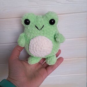 Op de afbeelding: Een groene gehaakte knuffel van een kikker met een witte buik en zwarte ogen. De kikker wordt in een hand vastgehouden.