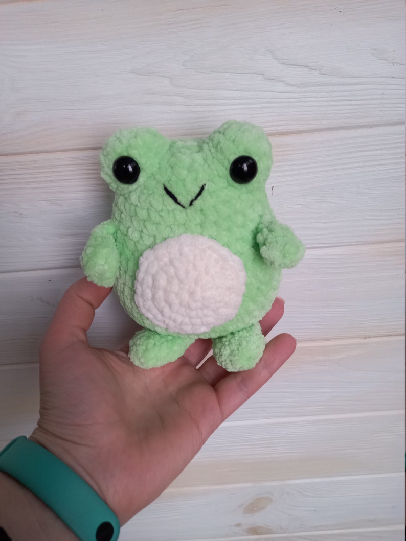 Op de afbeelding: Een groene gehaakte knuffel van een kikker met een witte buik en zwarte ogen. De kikker wordt in een hand vastgehouden.