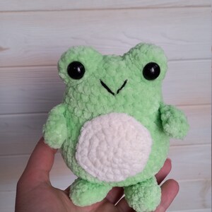Op de afbeelding: Een groene gehaakte knuffel van een kikker met witte gehaakte details en zwarte knoopjes voor ogen.