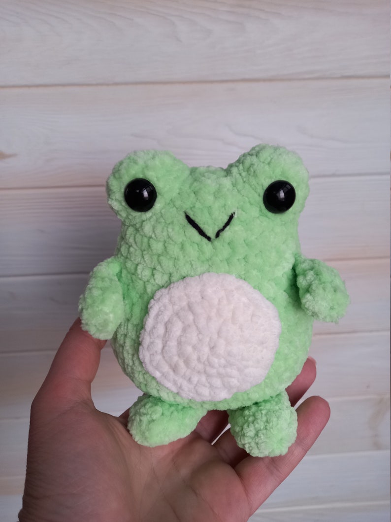Op de afbeelding: Een groene gehaakte knuffel van een kikker met witte gehaakte details en zwarte knoopjes voor ogen.