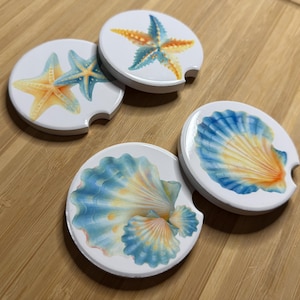 May include: Four white car coasters with ocean-themed designs. Two coasters feature starfish in shades of blue, orange, and yellow. The other two coasters display seashell designs in blue, orange, and yellow. Each coaster has a curved notch for easy removal.